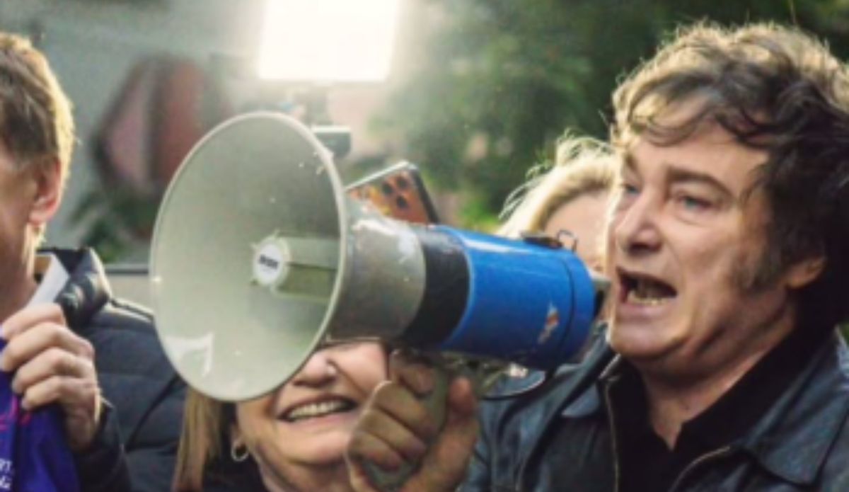 Javier Milei segura um megafone azul e fala em público durante um evento ao ar livre, cercado por outras pessoas que o acompanham e sorriem ao fundo.