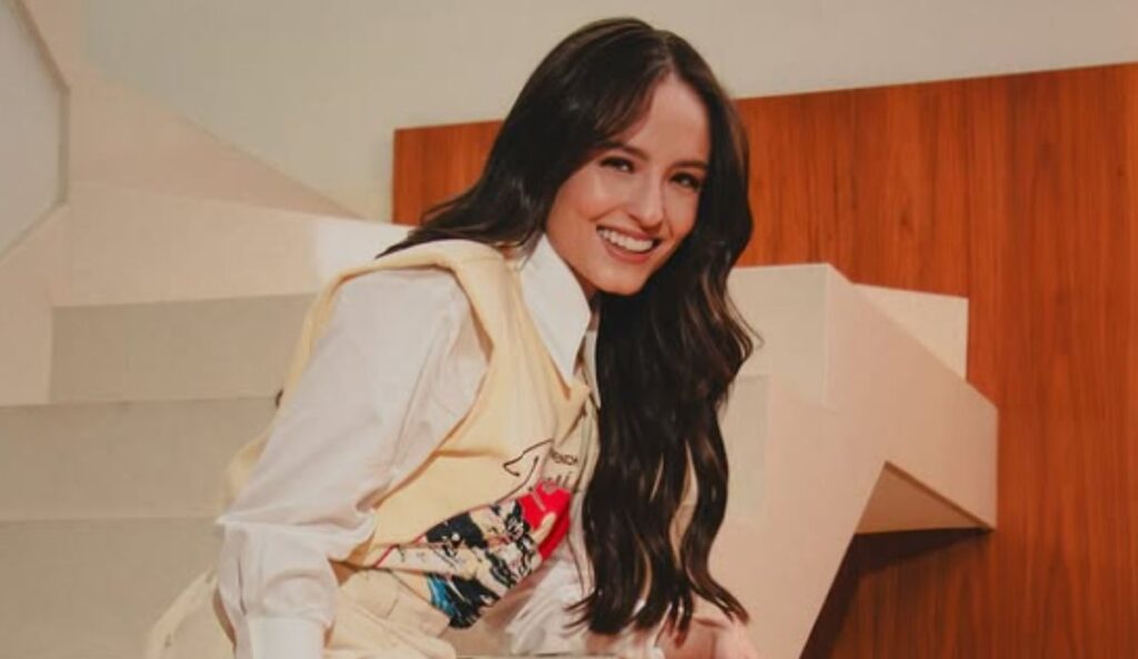 Larissa Manoela sentada em uma escada, sorrindo, usando camisa branca e colete claro, com fundo em tons de madeira.