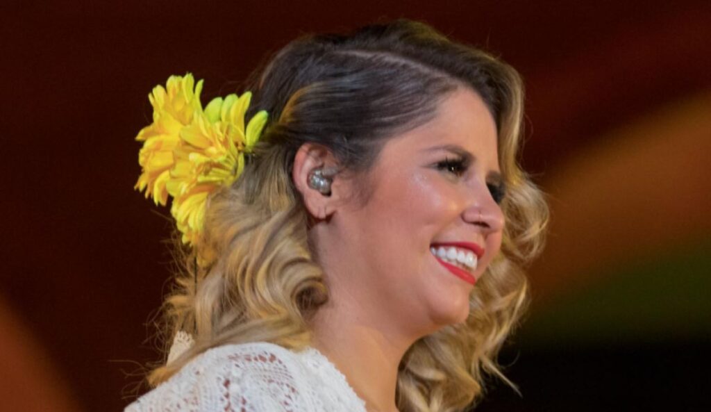 Marília Mendonça sorrindo de perfil, com cabelos loiros cacheados, usando um enfeite de flores amarelas no cabelo e batom vermelho, em um ambiente iluminado.