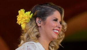 Marília Mendonça sorrindo de perfil, com cabelos loiros cacheados, usando um enfeite de flores amarelas no cabelo e batom vermelho, em um ambiente iluminado.