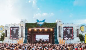 Palco principal do Nômade Festival em dia de show, com telões exibindo imagens em preto e branco, estrutura decorada com elementos naturais e céu azul ao fundo, enquanto uma multidão acompanha a apresentação