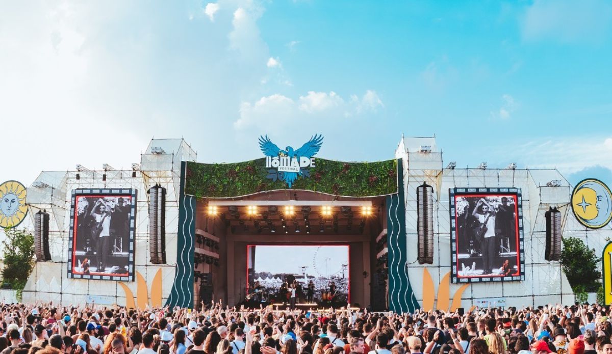 Palco principal do Nômade Festival em dia de show, com telões exibindo imagens em preto e branco, estrutura decorada com elementos naturais e céu azul ao fundo, enquanto uma multidão acompanha a apresentação