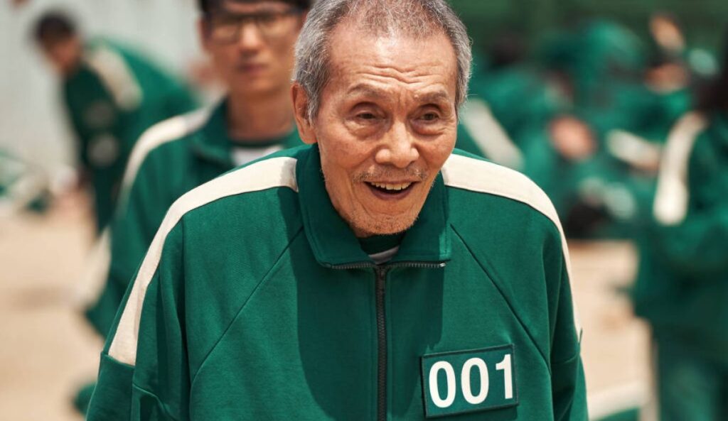 Oh Yeong-su em cena de Round 6, vestindo uniforme verde com o número 001, personagem marcante da série sul-coreana da Netflix.