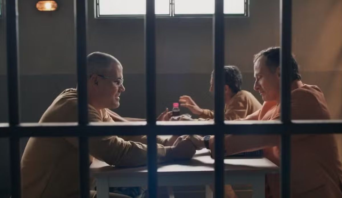 Cena de Três Graças mostra dois homens usando uniforme prisional conversando e segurando as mãos em uma mesa dentro da prisão, vistos através das grades.