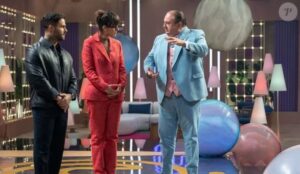 Três jurados do MasterChef Confeitaria 2025 conversam em um estúdio colorido; um deles usa terno azul claro, outro veste terno vermelho e o terceiro está com jaqueta preta. O cenário tem luzes suaves e grandes bolas decorativas no chão.