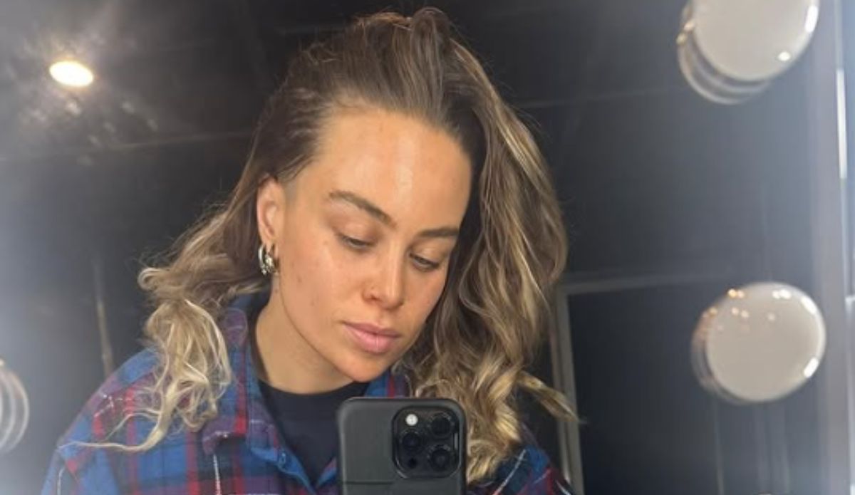 Tash Sultana posa em frente ao espelho com expressão serena, usando camisa xadrez azul e vermelha.