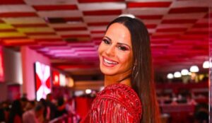 Viviane Araújo sorri para a câmera usando um vestido vermelho brilhante, com maquiagem combinando, em um ambiente iluminado com tons de vermelho e rosa ao fundo.
