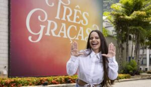 Viviane Araújo posa sorridente em frente ao painel da novela “Três Graças”, usando camisa branca e fazendo gesto com as mãos nos Estúdios Globo.