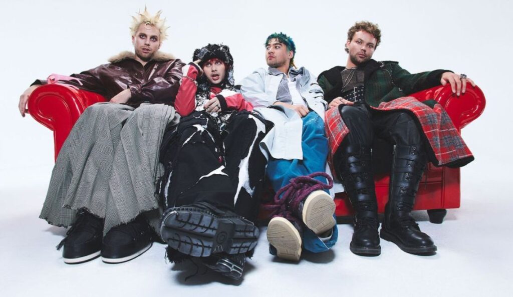 Integrantes do 5 Seconds of Summer sentados em um sofá vermelho, usando roupas em estilo alternativo e visual conceitual, em estúdio de fundo branco.