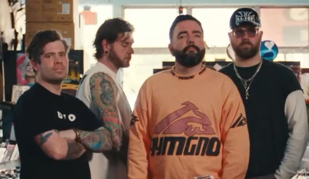 Os quatro integrantes do A Day To Remember posam dentro de uma loja de discos. Eles estão lado a lado, com expressão séria. O vocalista, ao centro, veste uma blusa oversized laranja; os demais membros usam roupas escuras, e um deles tem tatuagens visíveis nos braços. O ambiente ao fundo é iluminado e cheio de prateleiras com vinis e objetos diversos.