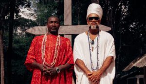 Carlinhos Brown e um líder marabaixeiro posam diante de uma grande cruz de madeira, usando vestes tradicionais e colares de contas, em cenário de floresta no Amapá.