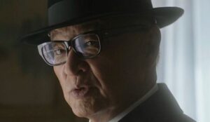 O ator Cary-Hiroyuki Tagawa usando chapéu e óculos em cena, com expressão séria e iluminação suave ao fundo.