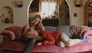 Cena de Margo’s Got Money Troubles, onde Michelle Pfeiffer e Elle Fanning estão sentadas em um sofá rosa. A mais velha está recostada e afaga o cabelo da mais jovem, que está deitada com a cabeça em seu colo. O ambiente é aconchegante, com almofadas estampadas e uma cozinha desfocada ao fundo.