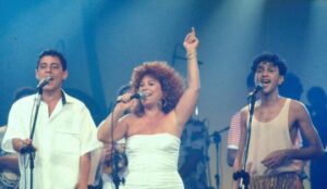 Chico Buarque, Beth Carvalho e Caetano Veloso cantando no palco durante um show de samba; ao centro, Beth de vestido branco com o braço levantado, acompanhada por dois Chico e Caetano que seguram microfones.
