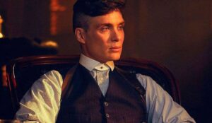 Cillian Murphy como Tommy Shelby em cena de “Peaky Blinders”. O personagem aparece sentado em um ambiente escuro, vestindo camisa social, colete e suspensórios, com expressão séria e iluminação dramática.