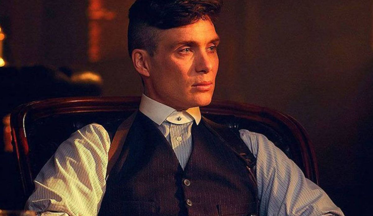 Cillian Murphy como Tommy Shelby em cena de “Peaky Blinders”. O personagem aparece sentado em um ambiente escuro, vestindo camisa social, colete e suspensórios, com expressão séria e iluminação dramática.