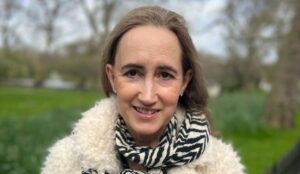 Sophie Kinsella sorrindo em área externa, usando cachecol estampado em preto e branco e casaco de lã claro; ao fundo, árvores desfocadas e gramado verde.