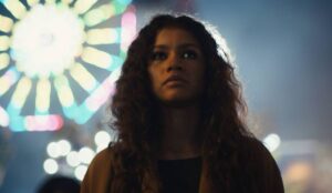 Zendaya em cena da 3ª temporada de “Euphoria”, com maquiagem marcada e expressão séria, diante de luzes coloridas ao fundo.