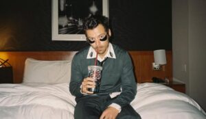 Harry Styles sentado em cama de hotel, usando patches nos olhos e segurando copo com canudo, em imagem associada à divulgação de “Aperture”.