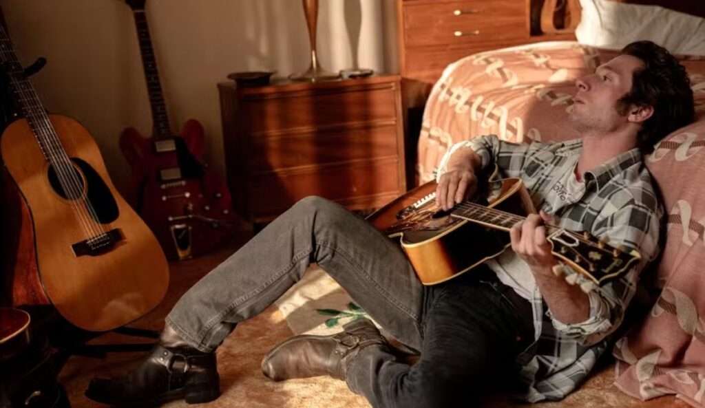 Cena de Springsteen: Salve-me do Desconhecido: músico deitado no chão do quarto toca violão cercado por guitarras, em momento íntimo de composição.