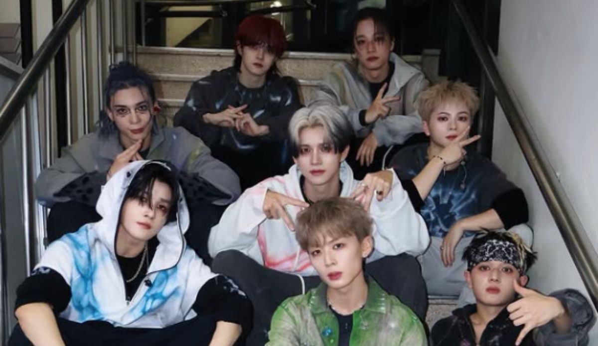 Grupo de j-pop ONE OR EIGHT posa em escadaria, com oito integrantes usando roupas em tons escuros e estética streetwear, em imagem promocional.