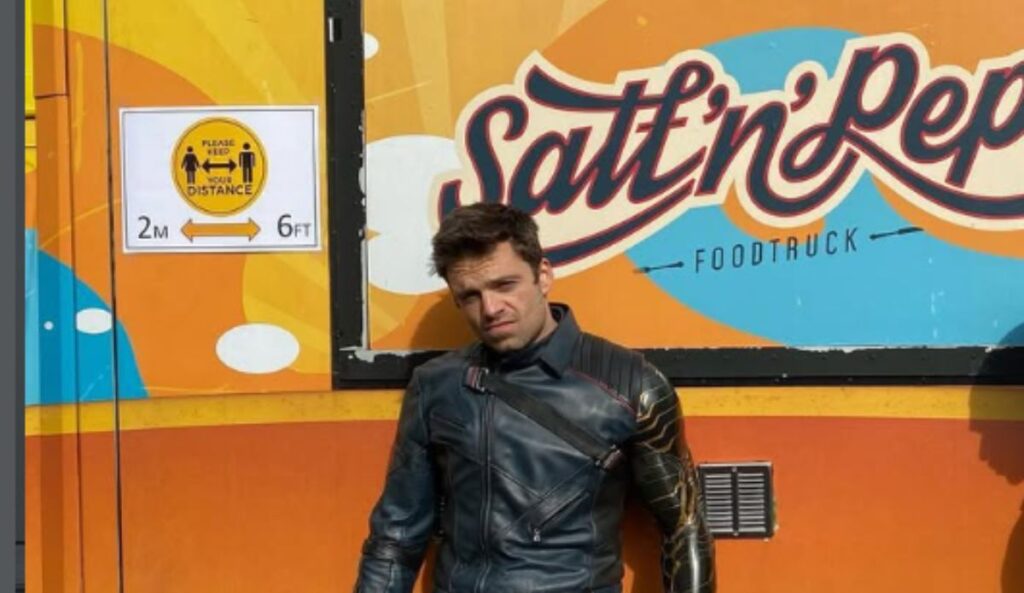 Sebastian Stan usando jaqueta de couro escura e braço metálico característico do Soldado Invernal, encostado em um food truck colorido ao ar livre.