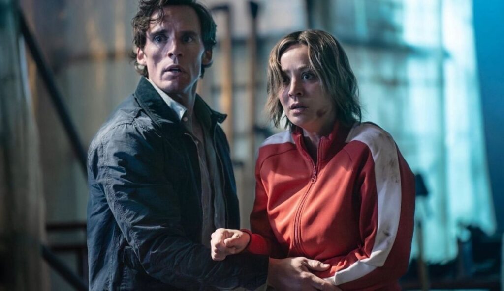 Kaley Cuoco e Sam Claflin em cena da série “Vanished”, em ambiente industrial escuro, com expressões de tensão.