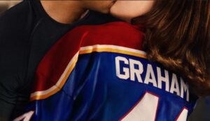 Casal protagonista de “Amores Improváveis” se beija enquanto veste uniforme de hóquei com nome Graham nas costas, série do Prime Video baseada nos livros de Elle Kennedy.