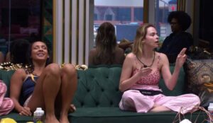 Ana Paula Renault conversa sentada no sofá da sala do BBB 26, gesticulando durante diálogo, enquanto Maxiane aparece ao lado rindo; ao fundo, outros participantes observam a cena.