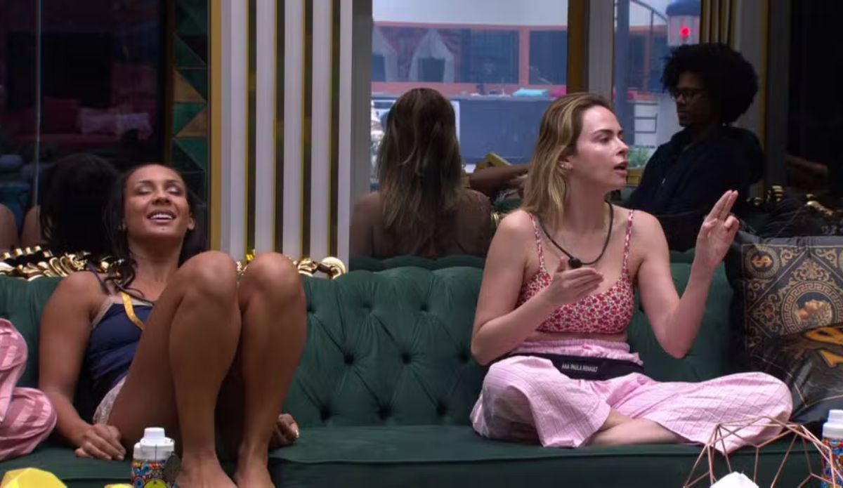 Ana Paula Renault conversa sentada no sofá da sala do BBB 26, gesticulando durante diálogo, enquanto Maxiane aparece ao lado rindo; ao fundo, outros participantes observam a cena.