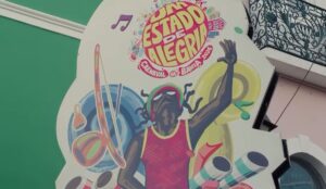 Painel artístico com ilustração de personagem negro celebrando o Carnaval, com a frase “Um Estado de Alegria – Carnaval da Bahia 2026” pintada em fachada de prédio histórico.
