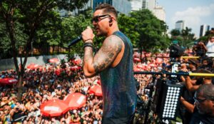 Felipe Pezzoni se apresenta em trio elétrico durante bloco de Carnaval em São Paulo, segurando o microfone enquanto uma multidão acompanha o desfile pelas ruas da cidade.
