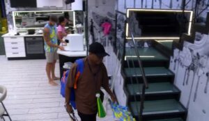 Edilson Capetinha com mochila nas costas dentro da casa do BBB 26, enquanto outros participantes circulam pela cozinha ao fundo.