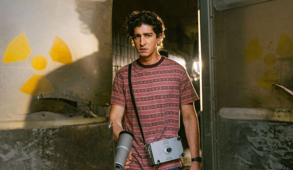 Johnny Massaro como seu personagem na série “Emergência Radioativa” em ambiente industrial deteriorado, segurando um medidor portátil de radiação; ao fundo, símbolos de material radioativo pintados na parede.