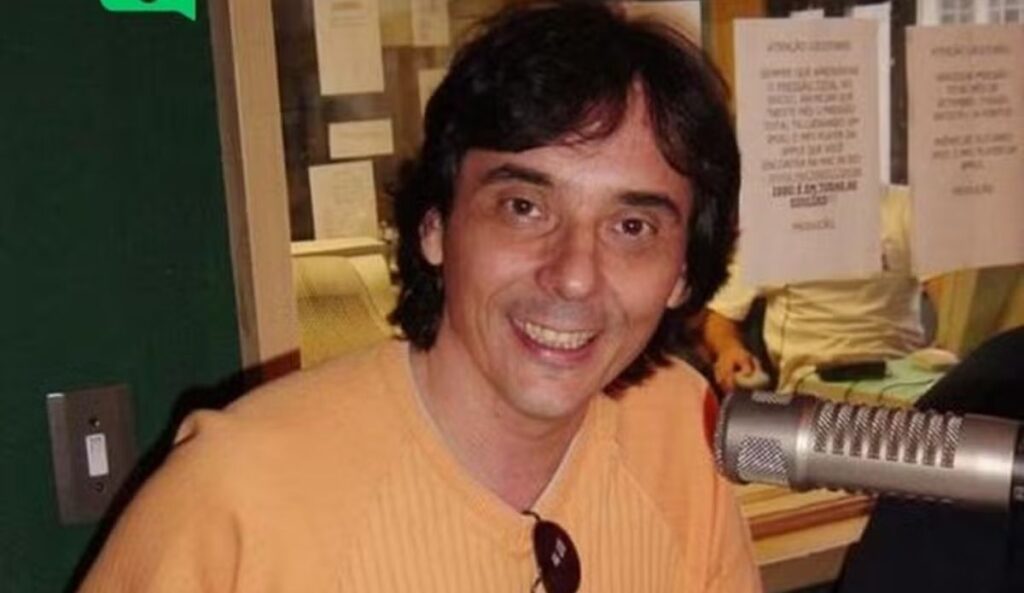 Ricardo Schnetzer sorri em estúdio, usando camiseta clara, sentado diante de um microfone profissional; ao fundo, há papéis fixados na parede.