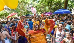 Foliões em bloco de rua do Carnaval no Rio de Janeiro, com multidão em parque arborizado e pessoa fantasiada de caranguejo.