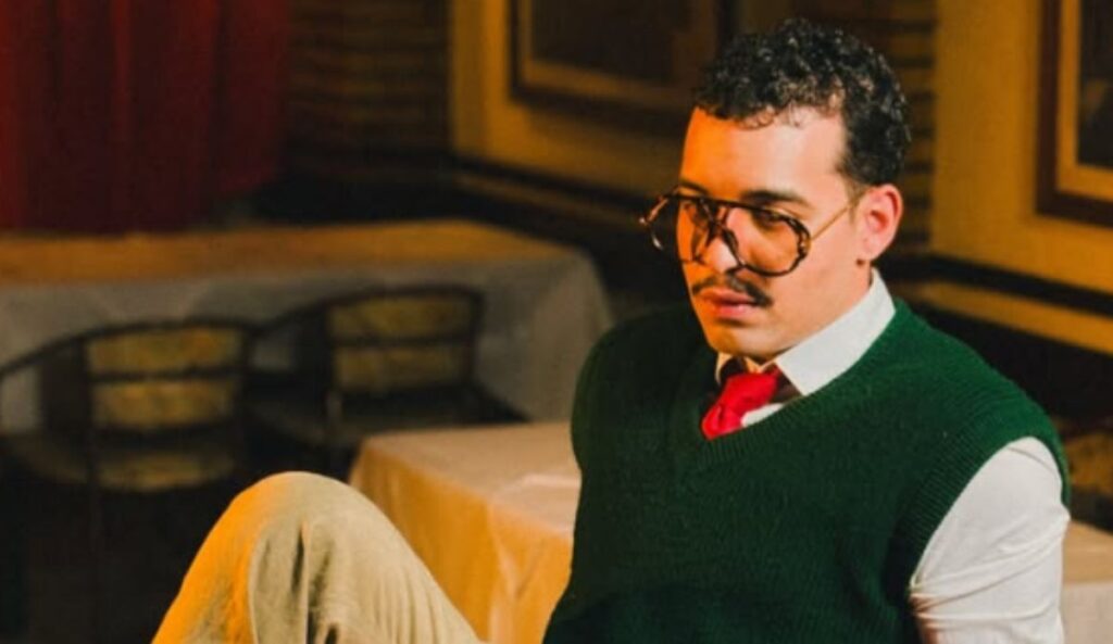 Cantor Bruno Leandro sentado em restaurante com luz amarelada, usando suéter verde e gravata vermelha, em cena do clipe “Roma”