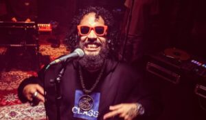 Rapper FBC canta ao microfone em ambiente de estúdio com iluminação vermelha, usando óculos escuros vermelhos e sorrindo; artista se apresenta no Lollapalooza Brasil 2026