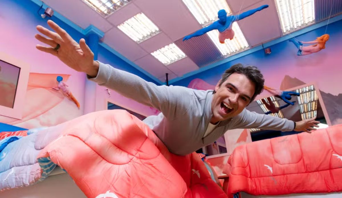 Tadeu Schmidt aparece sorrindo com os braços abertos em cenário colorido do Big Brother Brasil, com figuras suspensas no ar, em imagem promocional do BBB 26 usada para matéria sobre que horas começa o programa e a Prova do Líder.