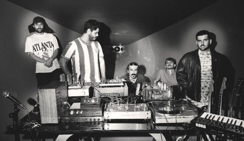 Integrantes da banda Jovem Dionísio em estúdio com equipamentos analógicos durante produção do álbum “Migalhas”, marcando nova fase.