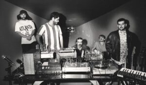 Integrantes da banda Jovem Dionísio em estúdio com equipamentos analógicos durante produção do álbum “Migalhas”, marcando nova fase.