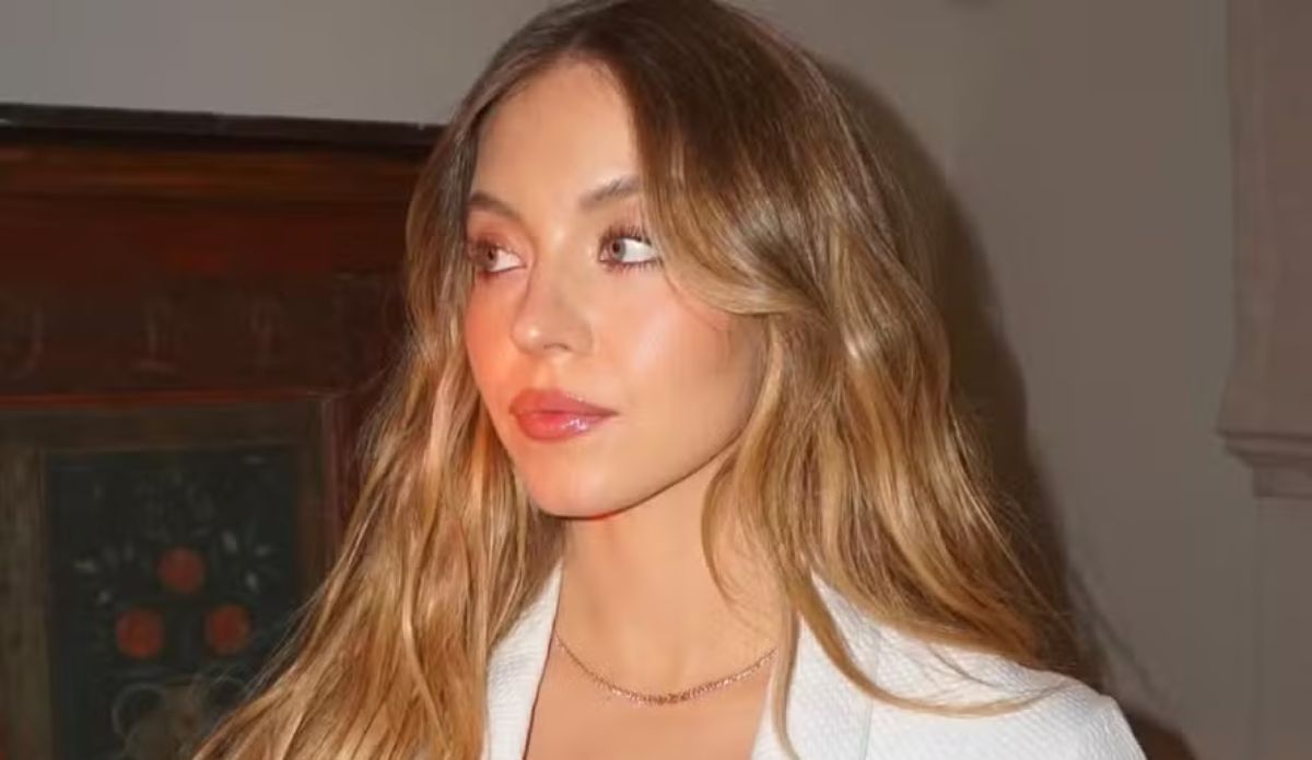 Sydney Sweeney em close com maquiagem natural e cabelo loiro ondulado, usando blazer branco; atriz teve cena cortada de "O Diabo Veste Prada 2"