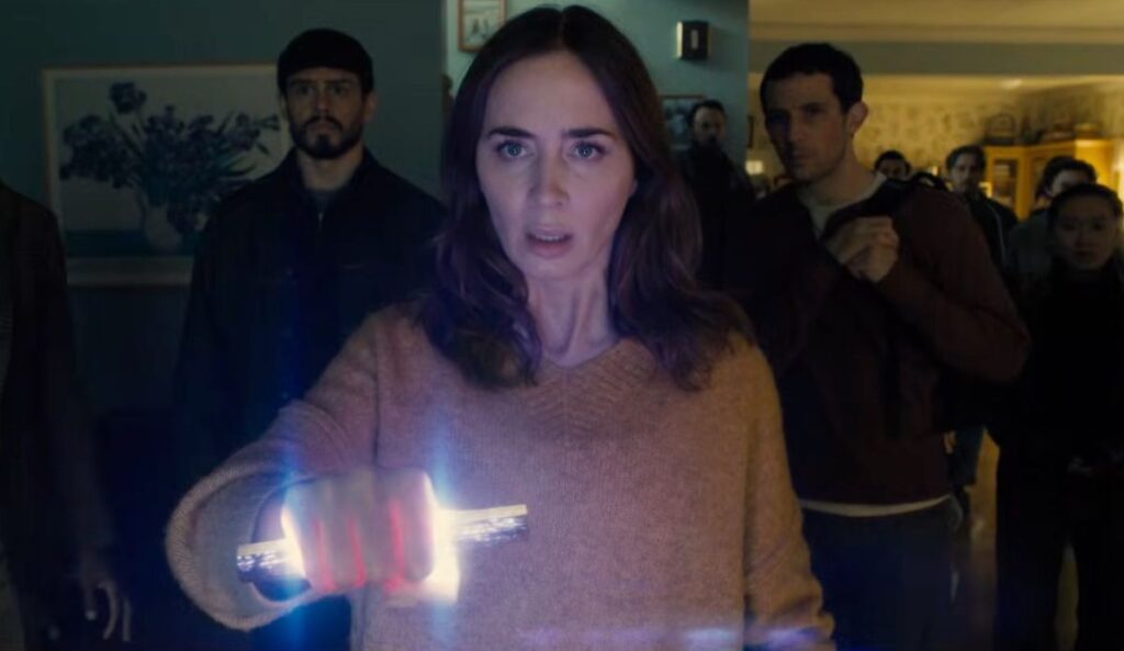 mily Blunt aparece assustada segurando um objeto luminoso em cena de “Dia D”, novo filme de Steven Spielberg. Ao fundo, várias pessoas observam a situação em uma sala escura.