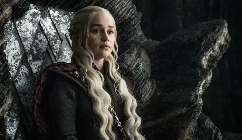 Emilia Clarke como Daenerys Targaryen, com cabelos platinados, sentada no trono de Pedra do Dragão em cena de Game of Thrones.