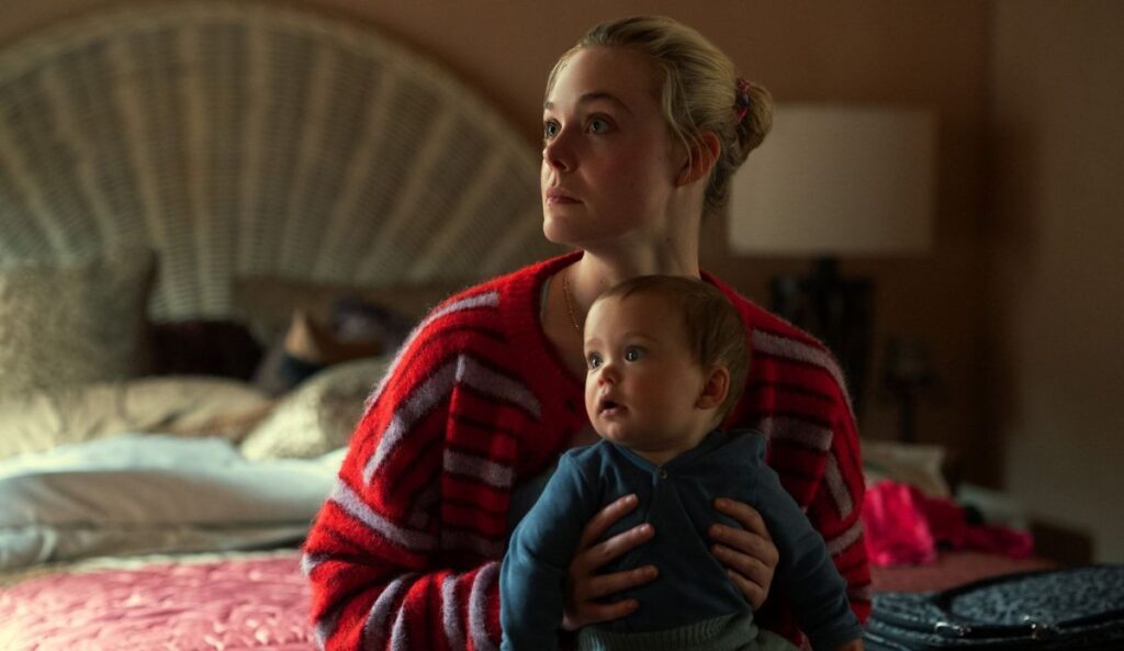 Elle Fanning aparece segurando um bebê no colo em cena da série “Margô Está em Apuros”, dentro de um quarto com iluminação baixa e expressão apreensiva.