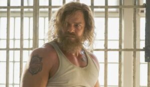 Alan Ritchson aparece com barba e cabelo comprido em cena de Motor City, diante de grades de uma prisão.