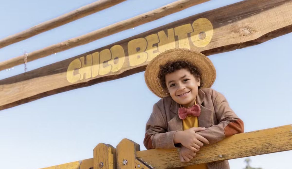 Isaac Amendoim aparece caracterizado como Chico Bento, sorrindo e usando chapéu de palha, apoiado em uma cerca de madeira