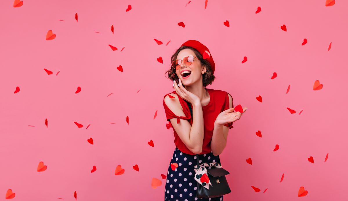 Mulher sorridente em cenário rosa com corações vermelhos para playlist de músicas do Dia dos Namorados.