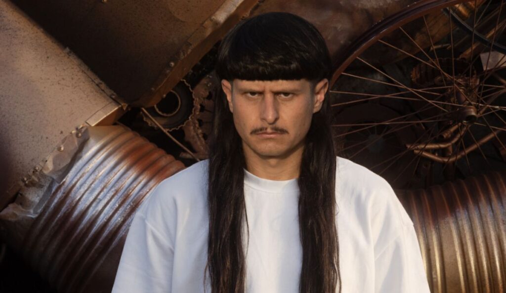Oliver Tree posa com expressão séria diante de uma estrutura metálica enferrujada. O artista usa uma camiseta branca larga e exibe seu visual característico, com franja reta, cabelo longo escuro e bigode fino. Ao fundo, aparecem rodas e peças metálicas em tons de cobre e ferrugem, reforçando a estética industrial da imagem.