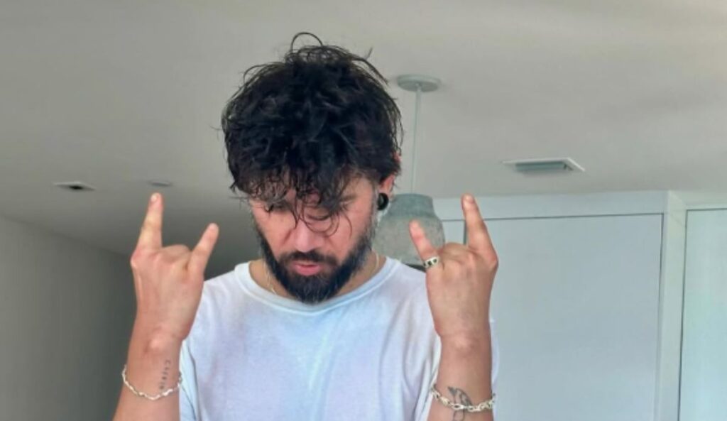 Skrillex aparece em um ambiente interno usando camiseta branca e cabelo molhado, fazendo o gesto de rock com as duas mãos. O produtor está de cabeça baixa, enquanto ao fundo é possível ver uma decoração minimalista com luminária suspensa.